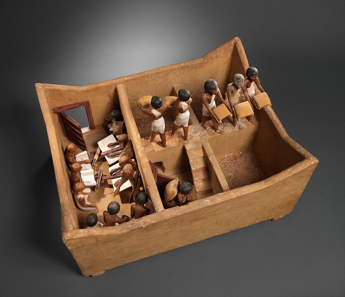 Ancient Egyptian wooden model with workers carrying grain and scribes recording supplies beside a storage area
